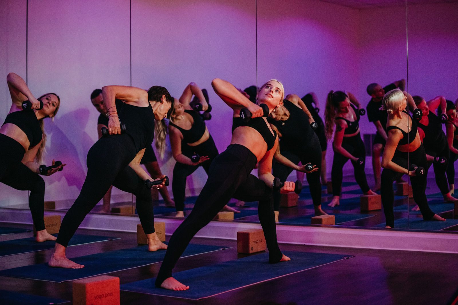 A yoga pod class in a side lunge with light hand weights under pink and purple studio light, reflected in the mirror