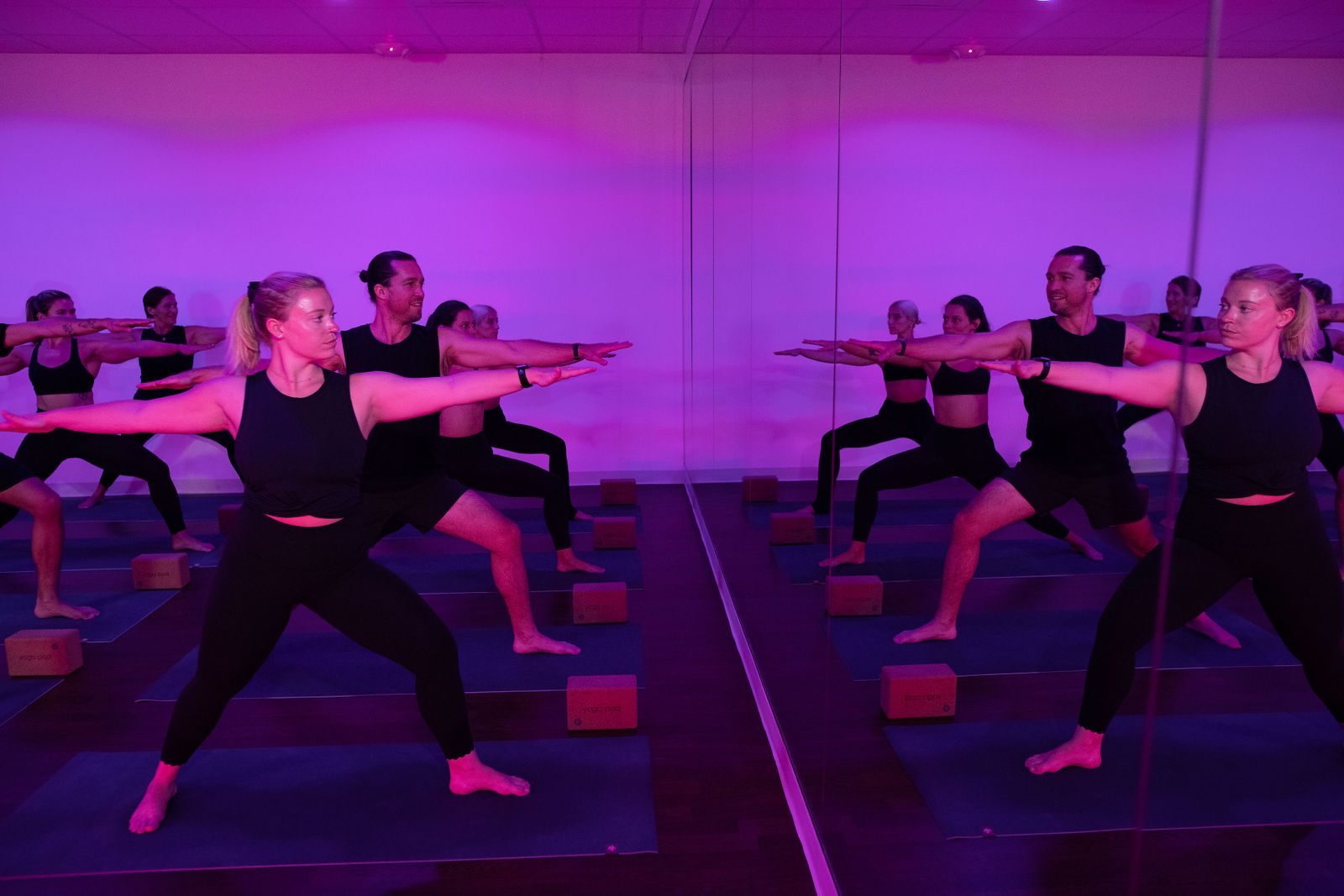 A yoga pod class holding Warrior 2 in pink studio light, reflected in the mirror
