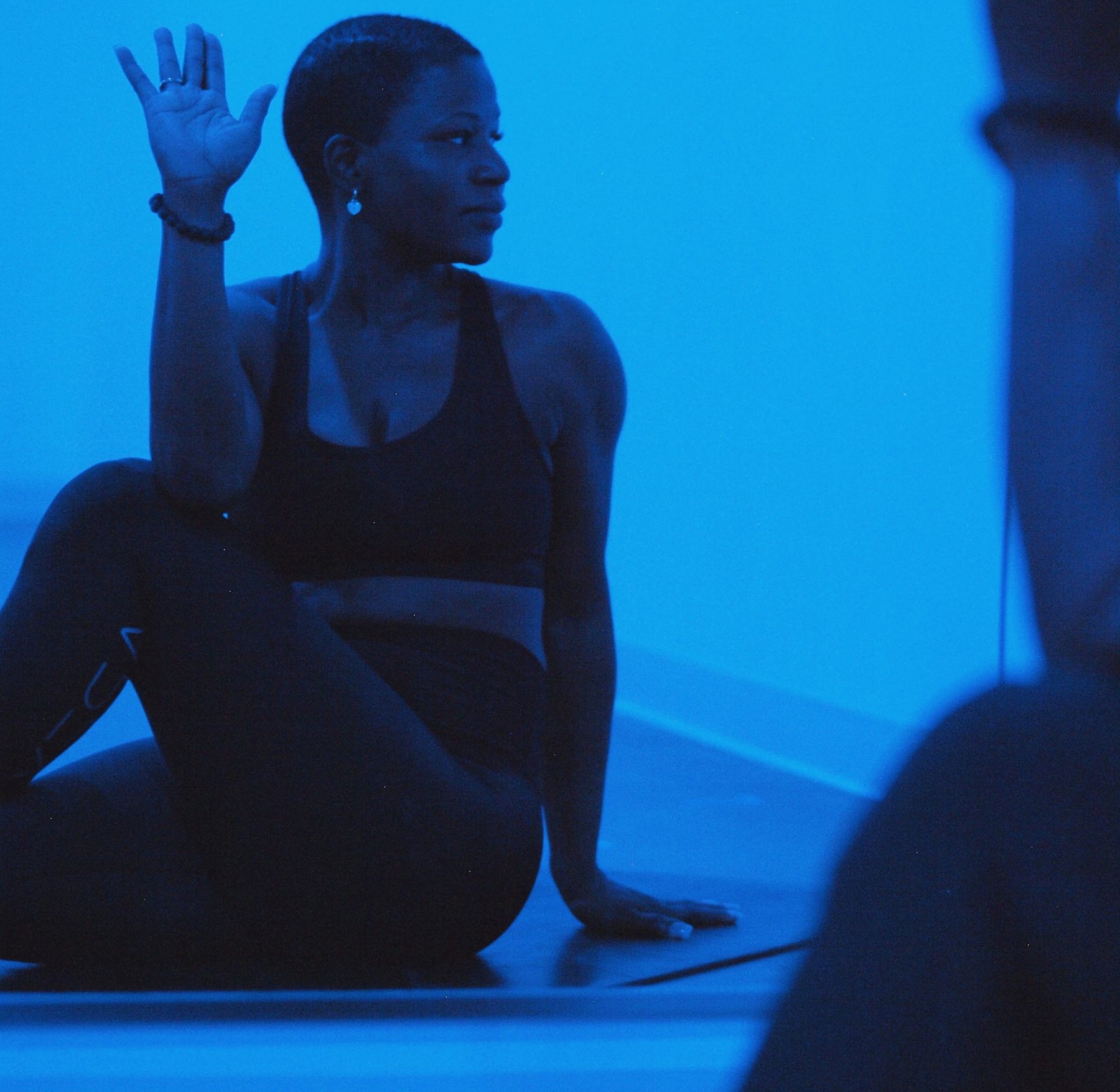 A student in a seated twist with one hand raised, glancing over her shoulder under blue studio light