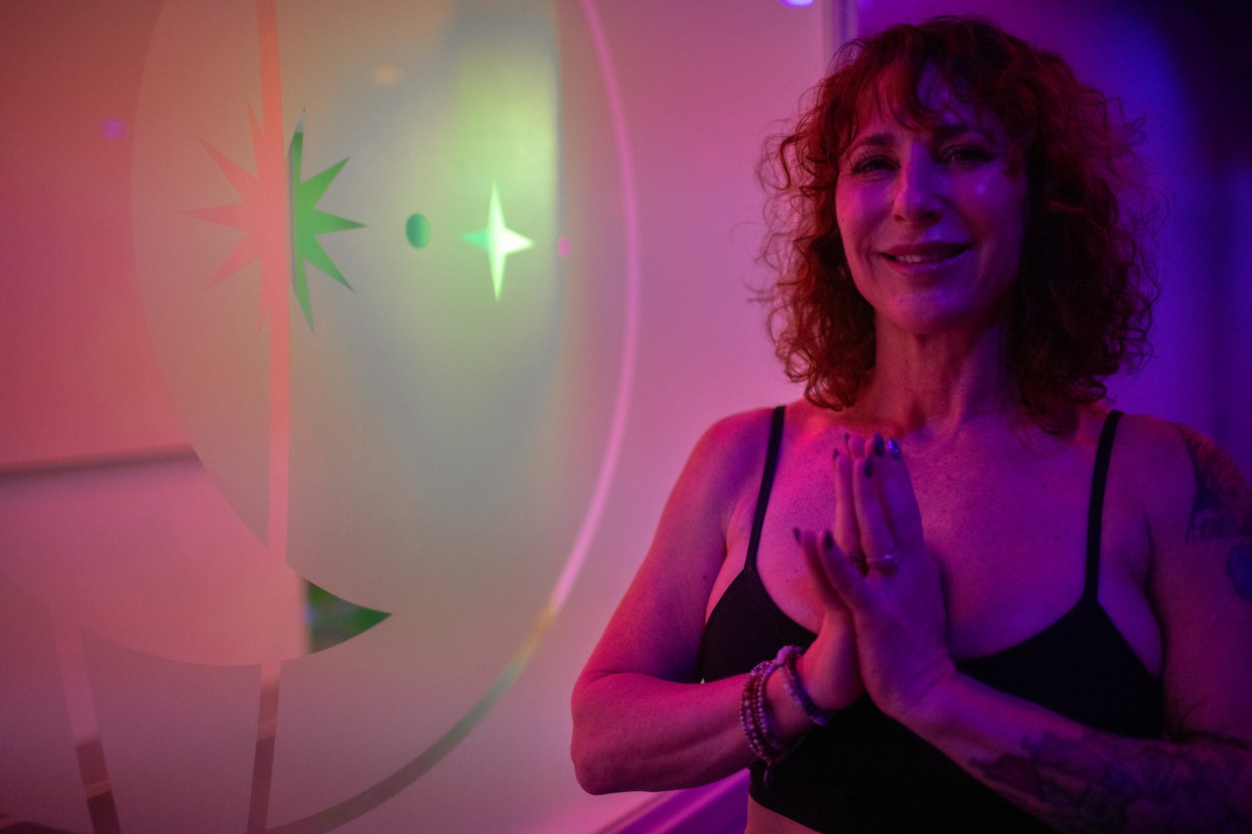 A smiling yoga pod student with her hands at heart center in glowing pink and purple studio light