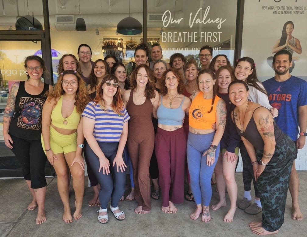 A smiling group of yoga pod teachers and trainees standing together outside the studio windows