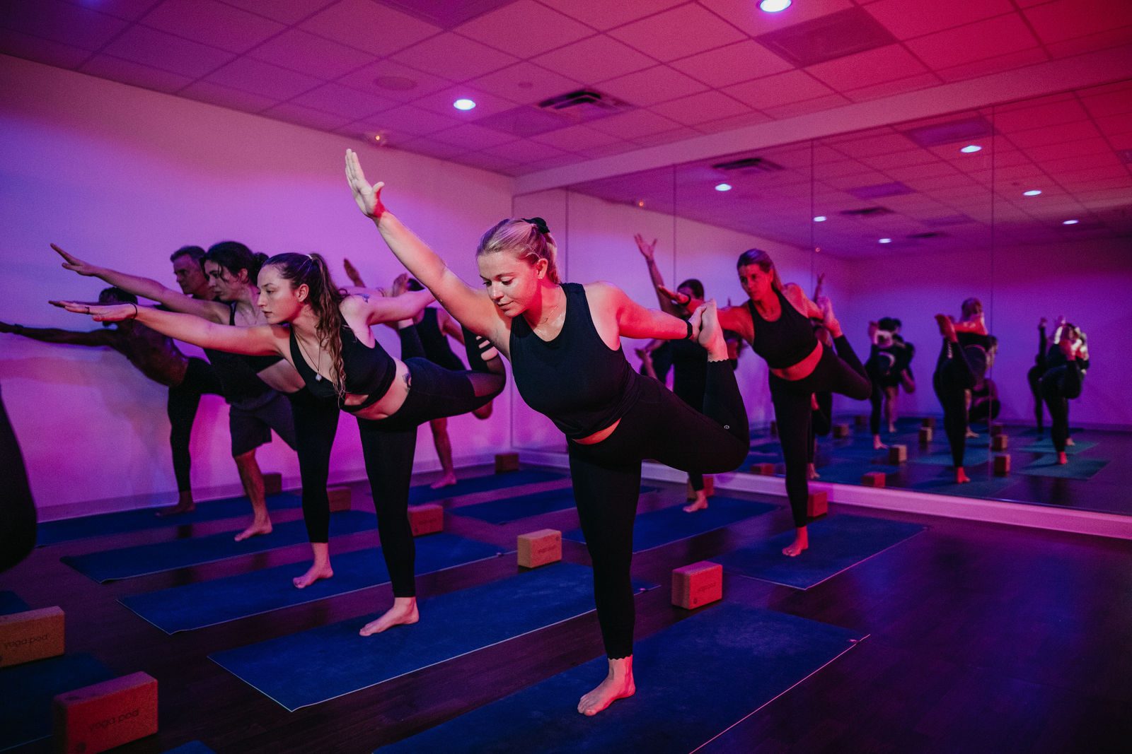 A yoga pod class balancing in Dancer pose under pink and purple studio light, reflected in the mirror