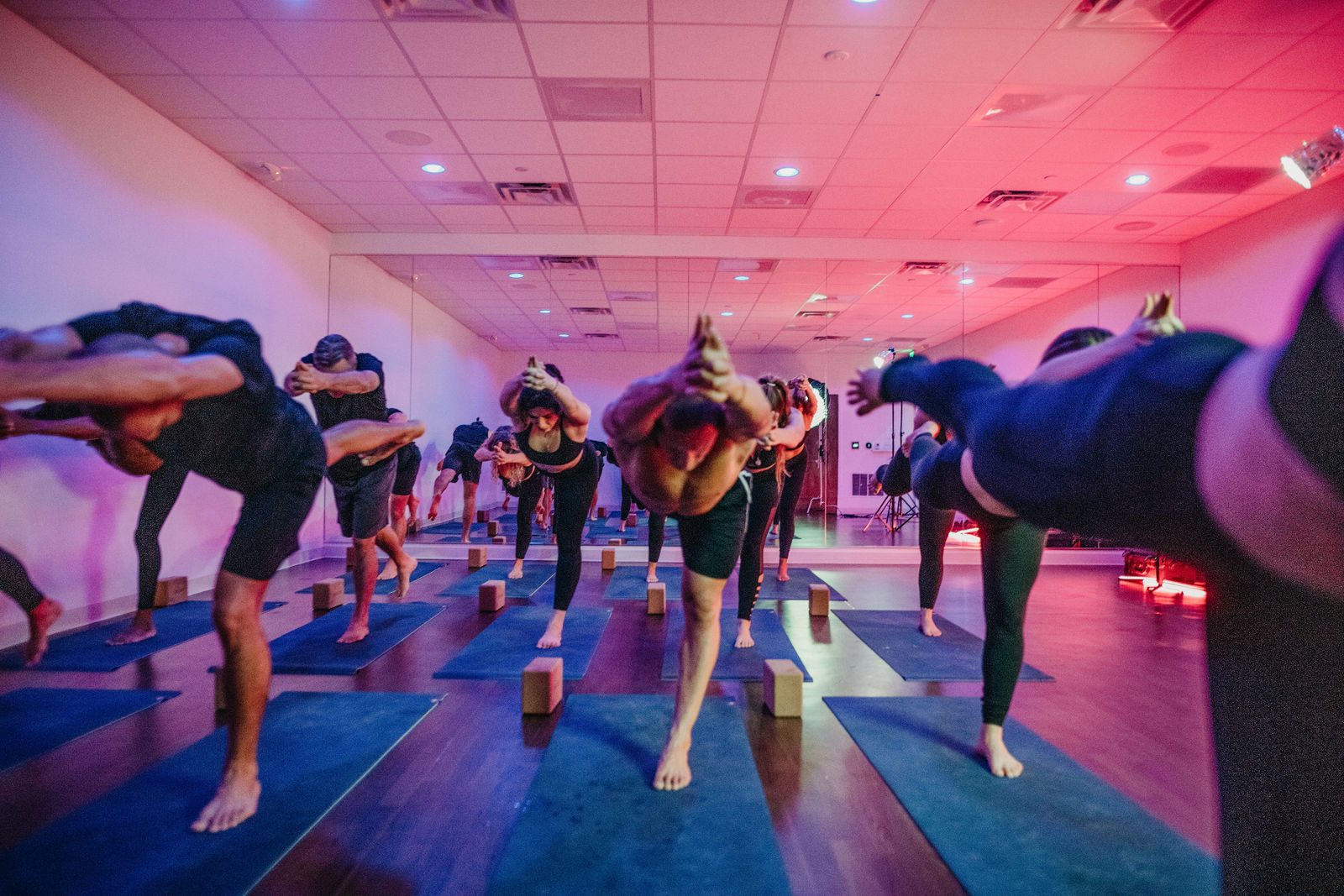 A full yoga pod class balancing in Warrior 3, facing the camera, under pink and purple studio light